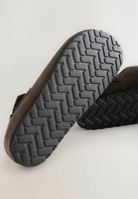 Braune Wildlederschlappen mit einer strukturierten grauen Gummisohle, die ein Zickzackmuster aufweist, und einem schwarzen Gummibezug.