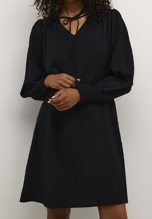 Kobieta ubrana w czarną sukienkę z długim rękawem z bufkami, marszczonymi mankietami i związanym dekoltem, stojąca na jednolitym tle.