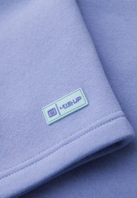 Tessuto di felpa lilla chiaro con texture liscia, con etichetta rettangolare mint con la scritta "LEVEL UP" in testo viola.