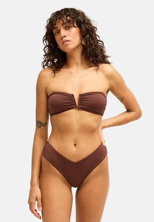 Femme aux cheveux bouclés et tatouages portant un haut de bikini marron bandeau et un bas de bikini taille haute sur un fond clair uni.