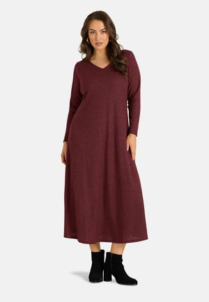 Femme portant une robe en maille bordeaux à manches longues et longueur cheville, avec des bottines noires, debout devant un fond clair uni.