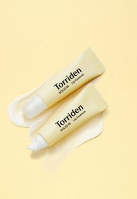 To gule tuber af Torriden læbeessens på en lys gul baggrund. Den ene tube har en rund spids, mens den anden har en spids applikator.