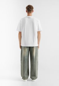 Witte katoenen t-shirt met korte mouwen, voorzien van een label aan de achterkant. Gepaard met groen-blauwe oversized denimbroek met een vervaagd textuur.