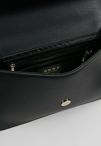 Svart läderhandväska med texturerad utsida, utrustad med guldknappslås, innerficka med dragkedja och synlig DKNY-märkesetikett inuti.