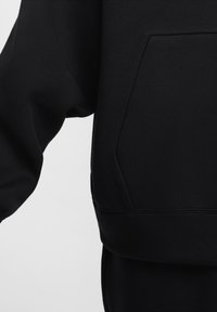 Černá mikina s měkkou texturou, volným střihem a přední kapsou ve tvaru klokánka s velkým zakřiveným tvarem blízko spodní části.