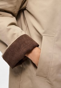 Μπεζ παλτό με καφέ κέντημα. Περιλαμβάνει πλαϊνή τσέπη και ομαλή υφή υφάσματος. Επικεντρώνεται στη χρηστικότητα και τα σχεδιαστικά στοιχεία.