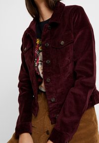 Bordeauxfarvet kordfløjlsjakke med knaplukning, fire forlommer og cropped design, kombineret med en grafisk T-shirt og brun kordfløjlsnederdel.