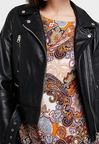 Chaqueta de moto de cuero negra con cremalleras y botones plateados, superpuesta a un vestido de vibrante estampado paisley en naranja, marrón y crema.