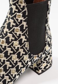 Gros plan d’une botte cheville noire et beige à motifs avec des détails en fil doré et un panneau latéral noir stretch.