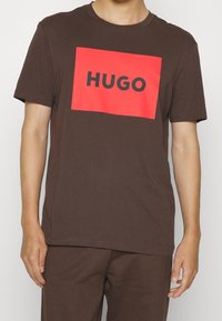 Hnědé bavlněné tričko s velkým červeným obdélníkovým potiskem s nápisem "HUGO" černě. Krátké rukávy, kulatý výstřih a volný střih.