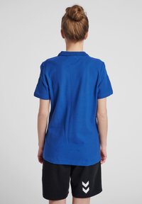 Blå pikétröja med krage, korta ärmar och en slät textur, matchad med svarta shorts med ett vitt chevronmönster.