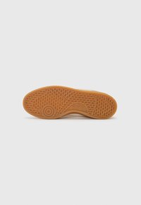 Sneaker zole ar teksturētu gumijas ārējo zoli. Gaiši brūna krāsa ar sarežģītu rakstu un aploku logotipu tuvu papēdim.