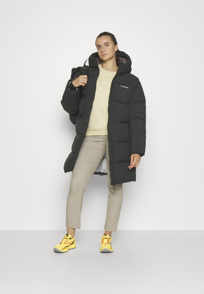 Frau steht in schwarzer Daunenjacke, beigen Hose und gelben Sneakers da und trägt einen schwarzen Rucksack über einer Schulter vor einem schlichten weißen Hintergrund.