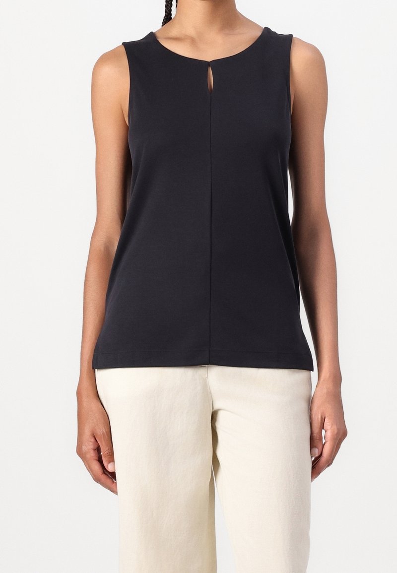Haut noir sans manches avec petit détail d'ouverture en forme de trou à l'avant, associé à un pantalon taille haute écru sur une personne debout.