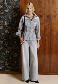 Blondynka w jasno szarej dopasowanej dżinsowej kurtce i szerokich spodniach, trzymająca czarną torebkę, stojąca na tle kwiecistych i drewnianych ścian.