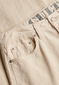 Détail d'un jean en denim beige montrant la poche avant, la boucle de ceinture, les rivets métalliques et un texte partiel en noir imprimé à l'intérieur de la ceinture.