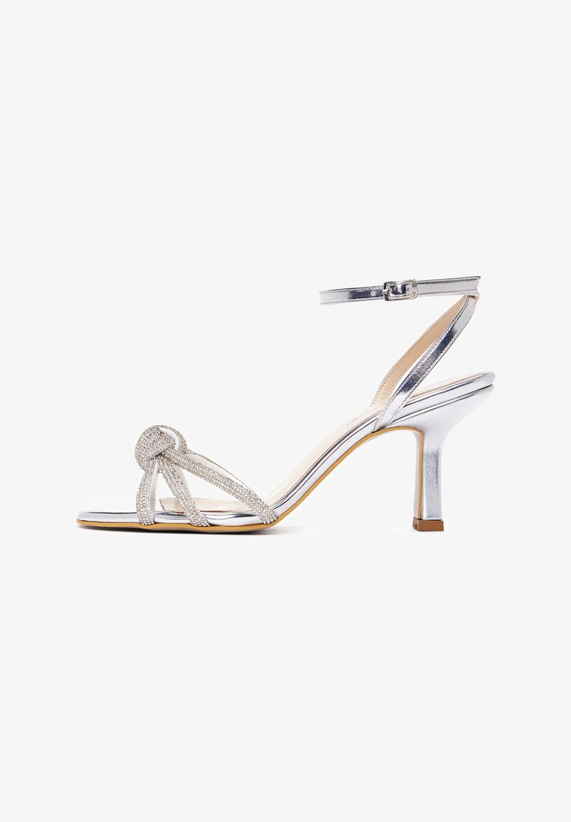Silver high-heel sandal with a metallic strap design, featuring a knot detail, ankle strap, and glossy finish.