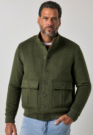 Homme barbu portant une veste verte boutonnée avec deux grandes poches frontales et un jean bleu clair, debout devant un fond uni.