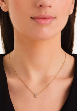 Collar de tono dorado con una cadena delgada y un colgante circular adornado con cristales transparentes, exhibido sobre un fondo neutro.