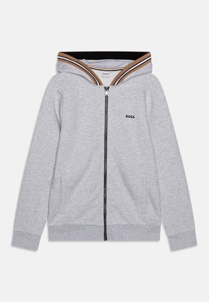 Haut à zip gris en tissu doux. Il arbore une capuche côtelée rayée noire et marron et un petit logo sur la poitrine gauche.