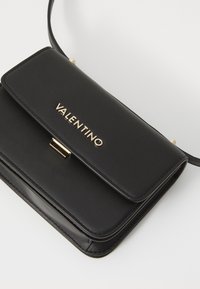 Borsa a tracolla in pelle nera con una chiusura dorata e la scritta "VALENTINO" impressa sulla patta. Forma rettangolare con design sottile e tracolla regolabile.