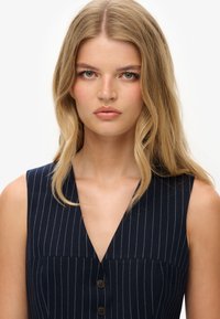 Granatowa sukienka bez rękawów z pionowymi białymi paskami, dekoltem w kształcie litery V i guzikami z przodu. Gładki materiał i dopasowany krój.