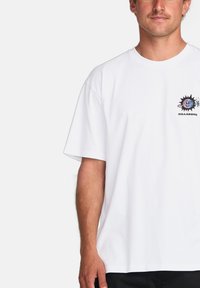 T-shirt en coton blanc à manches courtes et coupe décontractée. Présente un logo graphique coloré sur la poitrine gauche, représentant un design de soleil.