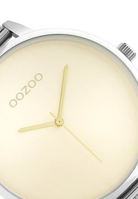Montre ronde avec un boîtier en acier inoxydable argenté, des aiguilles dorées et un cadran beige clair. La marque "OOZOO" est affichée en lettres discrètes.
