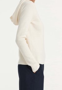 Felpa a coste di colore crema con tasca a marsupio e maniche lunghe, progettata con una vestibilità rilassata e una consistenza morbida, abbinata a pantaloni blu navy.