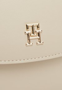 Krémová kožená kabelka s texturovaným povrchem, zdobená geometrickým logem v zlatém tónu a kontrastním šitím.