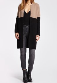 Knælang cardigan i beige og sort, lavet af blødt materiale. Åben front, ingen lukninger, med lange ærmer. Matchet med mørke skinny jeans og hælede støvler.