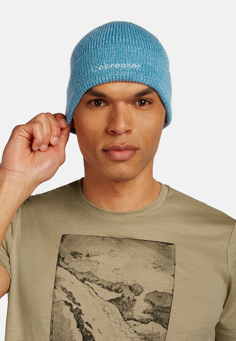 Beanie blu a maglia con texture a coste e un piccolo logo ricamato in bianco. La mano di un uomo aggiusta il cappello, indossando una maglietta grigia chiara sotto.