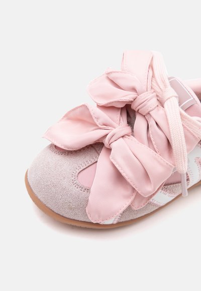 Gros plan sur une chaussure de bébé en suède rose avec de grands nœuds en satin noués dessus et des rayures blanches sur le côté.