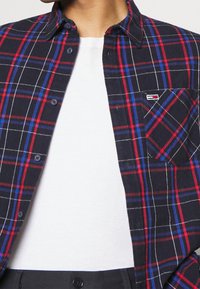 Koszulka w kratę z flaneli w kolorze granatowym, czerwonym i białym, z zapięciem na guziki oraz detalem kieszeni. Nosi się ją na białej koszulce.