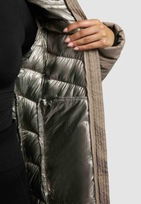 Mano che tiene aperto un cappotto beige, rivelando l'interno imbottito di un brillante colore argento metallico e una grande tasca interna.