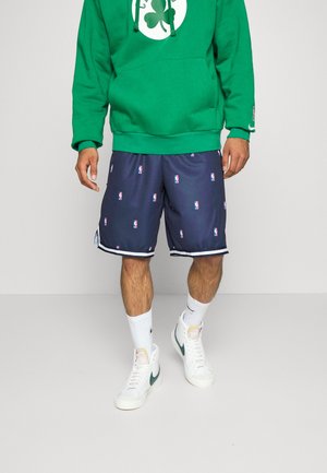 Mężczyzna w zielonej bluzie z kapturem z białym logo koniczyny, granatowych spodenkach NBA, białych skarpetkach i białych butach Nike z zielonym znakiem.