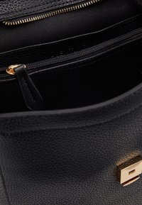 Sac à main en cuir noir avec une surface texturée, une fermeture éclair dorée, et un logo sur le devant ; l'intérieur comprend une poche zippée.