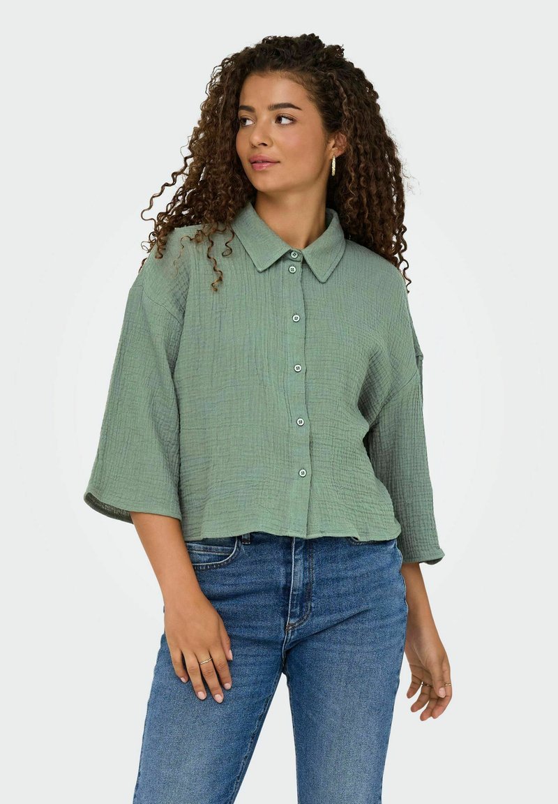Camicia cropped con bottoni in tessuto verde chiaro testurizzato, colletto a punta e maniche a tre quarti, abbinata a jeans in denim blu.