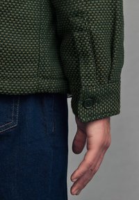 Grüne karierte Texturjacke mit einem Knopfmanschette, kombiniert mit dunkelblauen Jeans. Der Stoff zeigt ein gewebtes Muster.