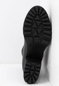 Semelle de bottes noires avec un motif de crampons épais, montrant des rainures triangulaires et rectangulaires, positionnée contre un angle de mur et de sol blanc.