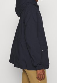 Námořnicky modrá mikina s kapucí, volného střihu, elastickými manžetami a boční kapsou se zapínáním na patent. Hladká textura a lehký materiál.