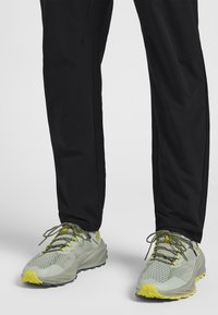 Baskets avec une tige en mesh gris clair, accents jaunes et semelle en caoutchouc texturé. Associées à un pantalon noir. Les lacets sont jaunes et verts.