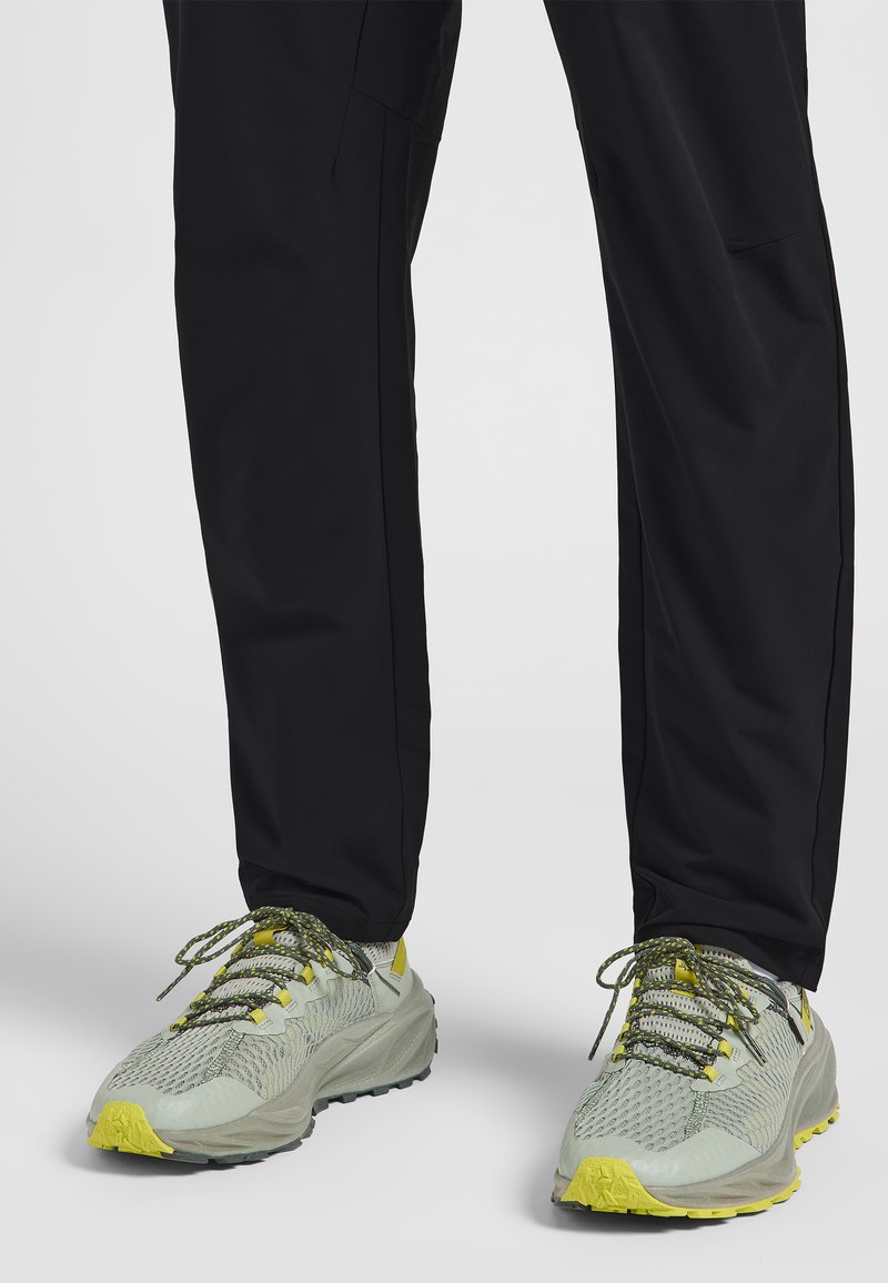 Baskets avec une tige en mesh gris clair, accents jaunes et semelle en caoutchouc texturé. Associées à un pantalon noir. Les lacets sont jaunes et verts.