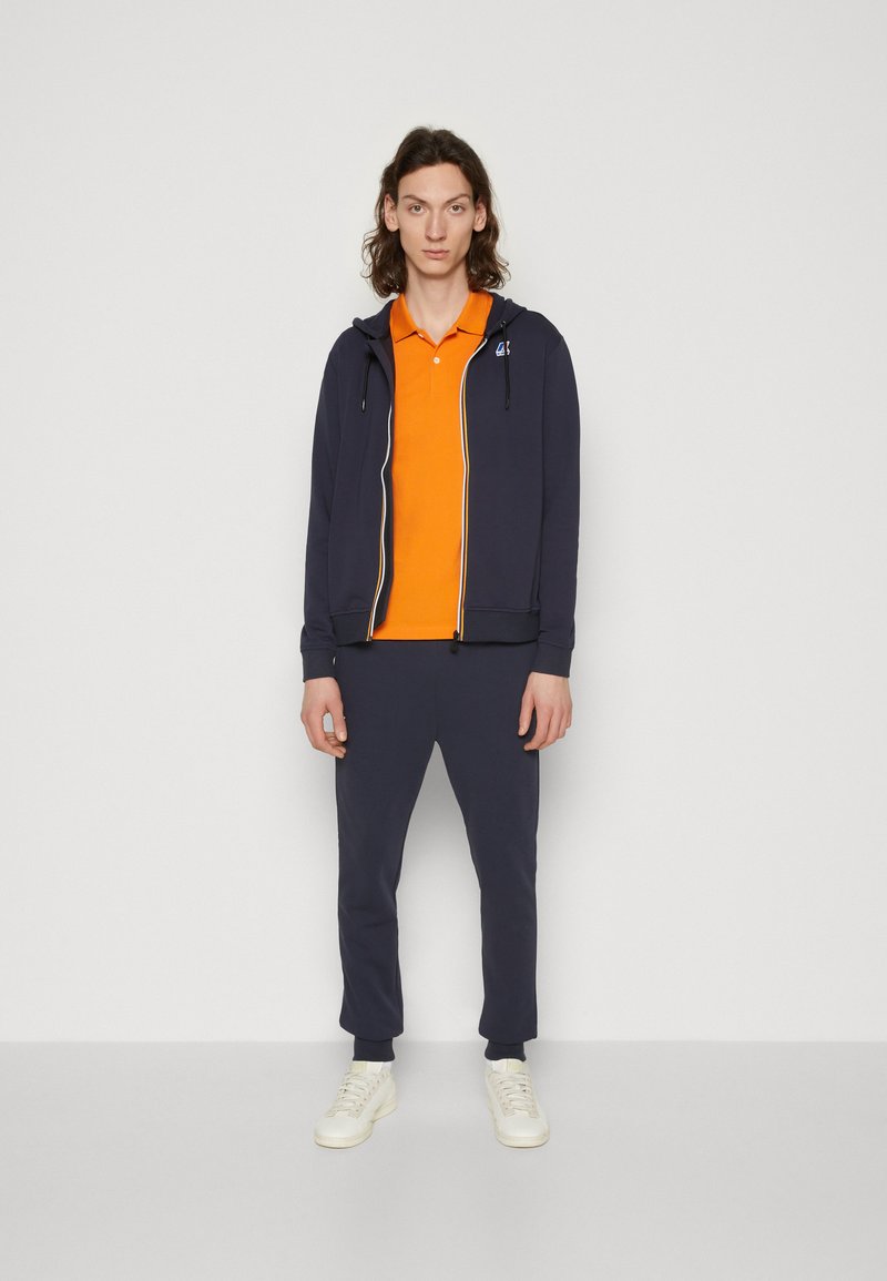 Marineblaue Zip-Hoodie mit orangefarbenem Poloshirt darunter und passenden marineblauen Jogginghosen. Weiße Schuhe. Glattes Material mit kontrastierenden Reißverschlussdetails.