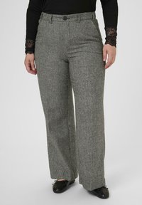 Žena v šedých herringbone širokých nohaviciach, čiernom topu s čipkovými rukávmi a čiernych baletných topánkach stojí na jednoduchom pozadí.