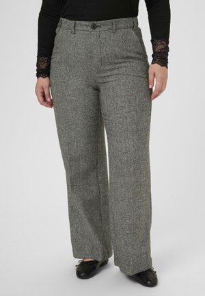 Frau in grauen Fischgrät-Hose mit weitem Bein, schwarzem Oberteil mit Spitzenärmeln und schwarzen Ballettschuhen, die vor einem schlichten Hintergrund steht.