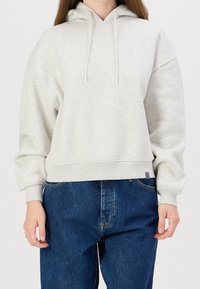 Světle šedá mikina s kapucí na stažení, žebrovaným lemem a volným střihem, doplněná tmavě modrými džínami. Materiál působí měkce a texturovaně.