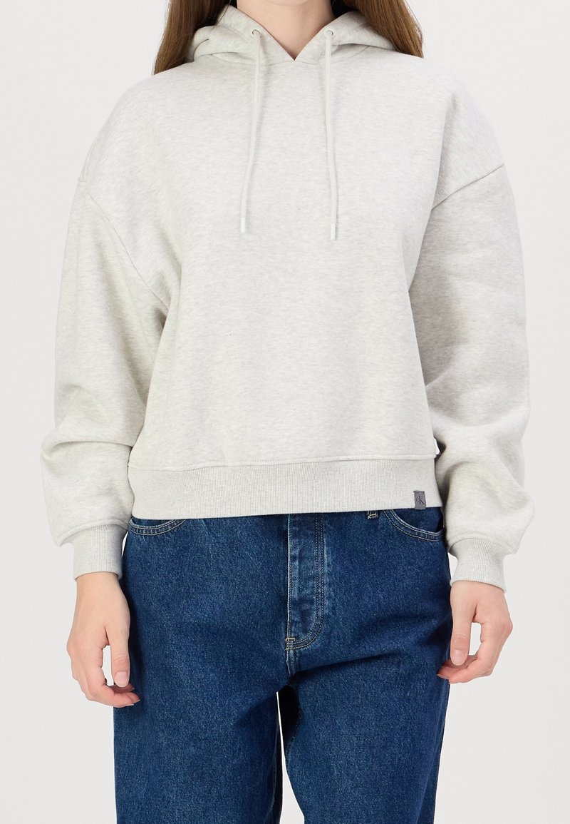Světle šedá mikina s kapucí na stažení, žebrovaným lemem a volným střihem, doplněná tmavě modrými džínami. Materiál působí měkce a texturovaně.