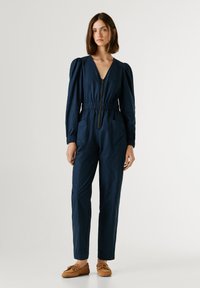 Ναυτικό ολόσωμο με μακριά φουσκωτά μανίκια, βαθύ V-λαιμόκοψη, ελαστική μέση, μπροστινό φερμουάρ και δύο μπροστινές τσέπες. Φορεμένο με ανοιχτό καφέ παπούτσια.