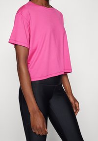 Kvinna klädd i en ljusrosa kortärmad topp och hög midja svarta leggings, som står mot en enfärgad ljus bakgrund.